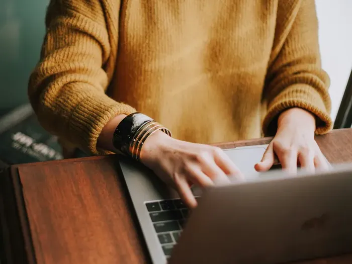 Close-up of person typing on a laptop wearing a cozy mustard sweater and bracelet at a wooden desk
